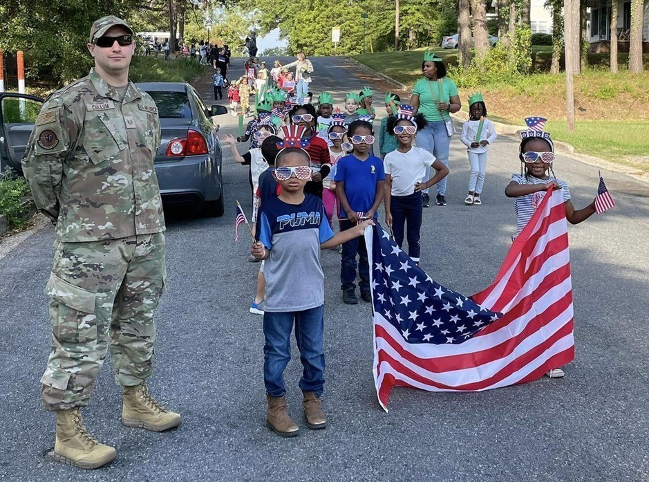 Kindergarten and 1st graders stand on the road with an American flag.